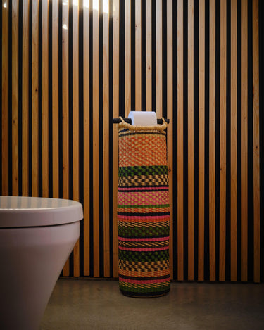 A handwoven tubular toilet roll holder with pink and green gingham design, placed against a striped wall.