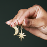 Hand holding gold earrings with star and moon designs against a dark background