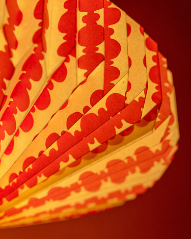 A close-up of an AARVEN pleated paper light shade with a repeating pink pattern on a warm white background, set against a solid dark red background.