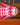 A close-up of AARVEN Pink Beads hanging paper light shade with a bold pink and white geometric pattern, shown against a softly blurred background with warm orange and brown tones.