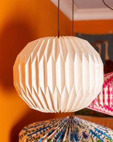 AARVEN white Sun Mask pleated paper pendant light hanging in a warm orange room, with other patterned ligh shades blurred in the background.