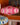 An AARVEN hanging paper light shade with a pink and white geometric pattern, suspended indoors against an orange wall with a framed artwork in the background.