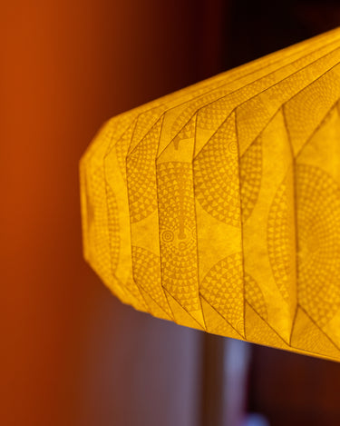 A close-up of a warm white paper lantern by AARVEN with a textured geometric Sun Mask pattern, shown against a blurred orange and dark brown background.