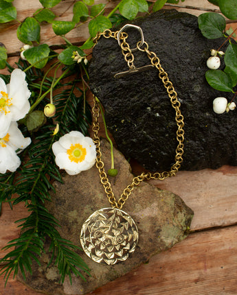 Gold necklace with a decorative sun mask pendant on a natural background with rocks and flowers