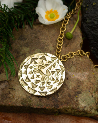 Gold necklace with a round, intricately designed pendant on a stone surface with a white flower.
