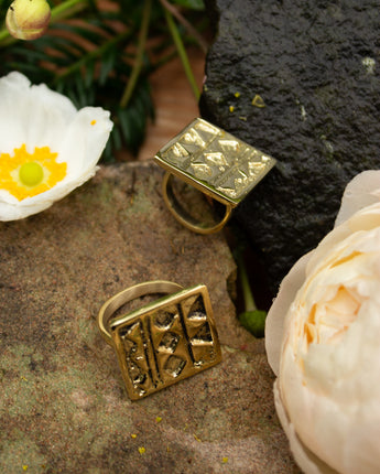 Two gold rings with geometric designs on a stone surface with flowers.