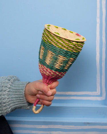 Hand holding a colourful woven rattle against a blue wall.