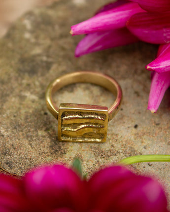 Gold ring with a square design on a stone surface with pink flowers.