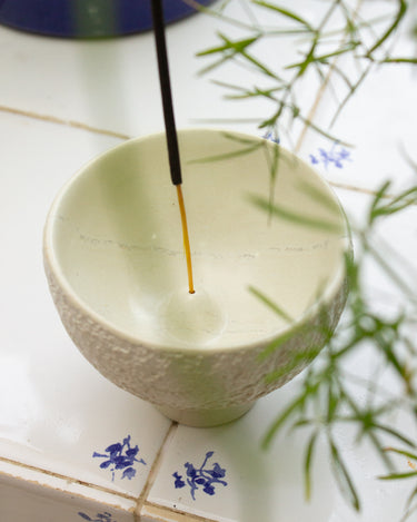 Handmade white soapstone incense holder with an AARVEN incense stick on a tiled surface, with a plant in the background.