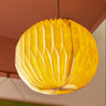 AARVEN large, pleated paper Sun Mask design pendant light shade in a warm off-white color, suspended from a black ceiling mount against a peach-coloured ceiling.