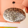A modern paper pendant light with a black and white sun mask pattern, suspended from a black ceiling mount against a peach-colored ceiling.