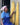 Woman in blue dress and headscarf holding yellow fibers against a gray wall.